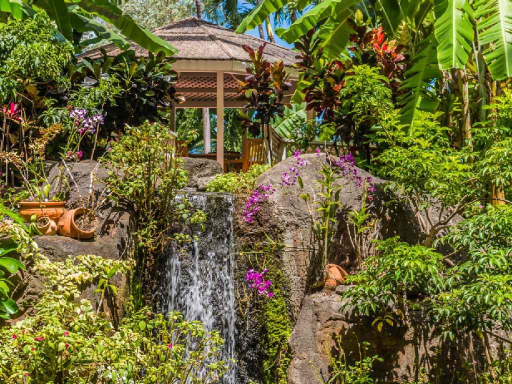 Un jardín tropical con una cascada, un pabellón y abundante vegetación.