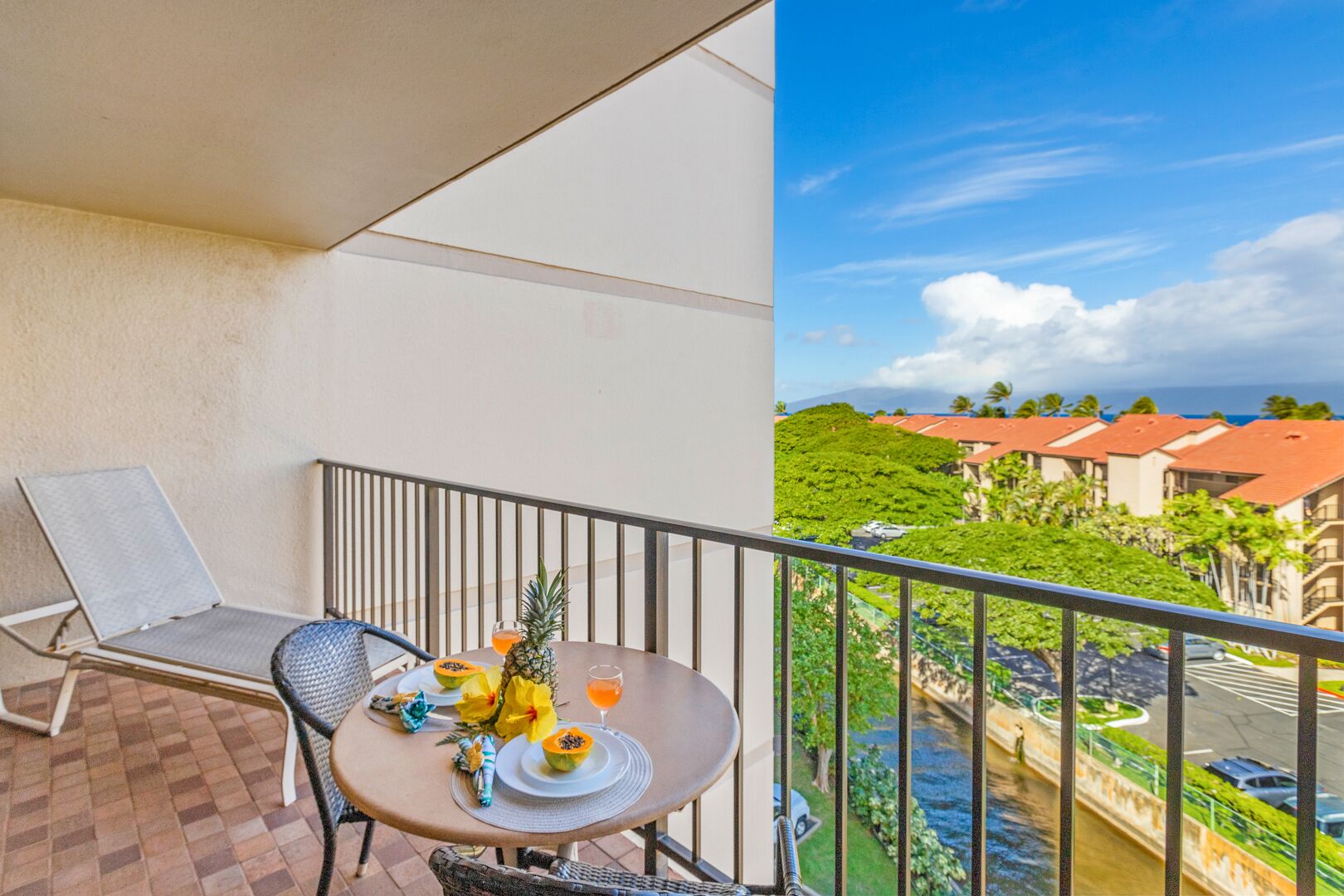 Un balcon avec une table dressée pour deux personnes, agrémentée de fruits tropicaux et d’une boisson, offrant une vue sur un paysage verdoyant et luxuriant, avec des bâtiments et un ciel dégagé.