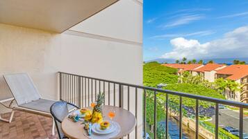 Un balcon avec une table dressée pour deux personnes, agrémentée de fruits tropicaux et d’une boisson, offrant une vue sur un paysage verdoyant et luxuriant, avec des bâtiments et un ciel dégagé.