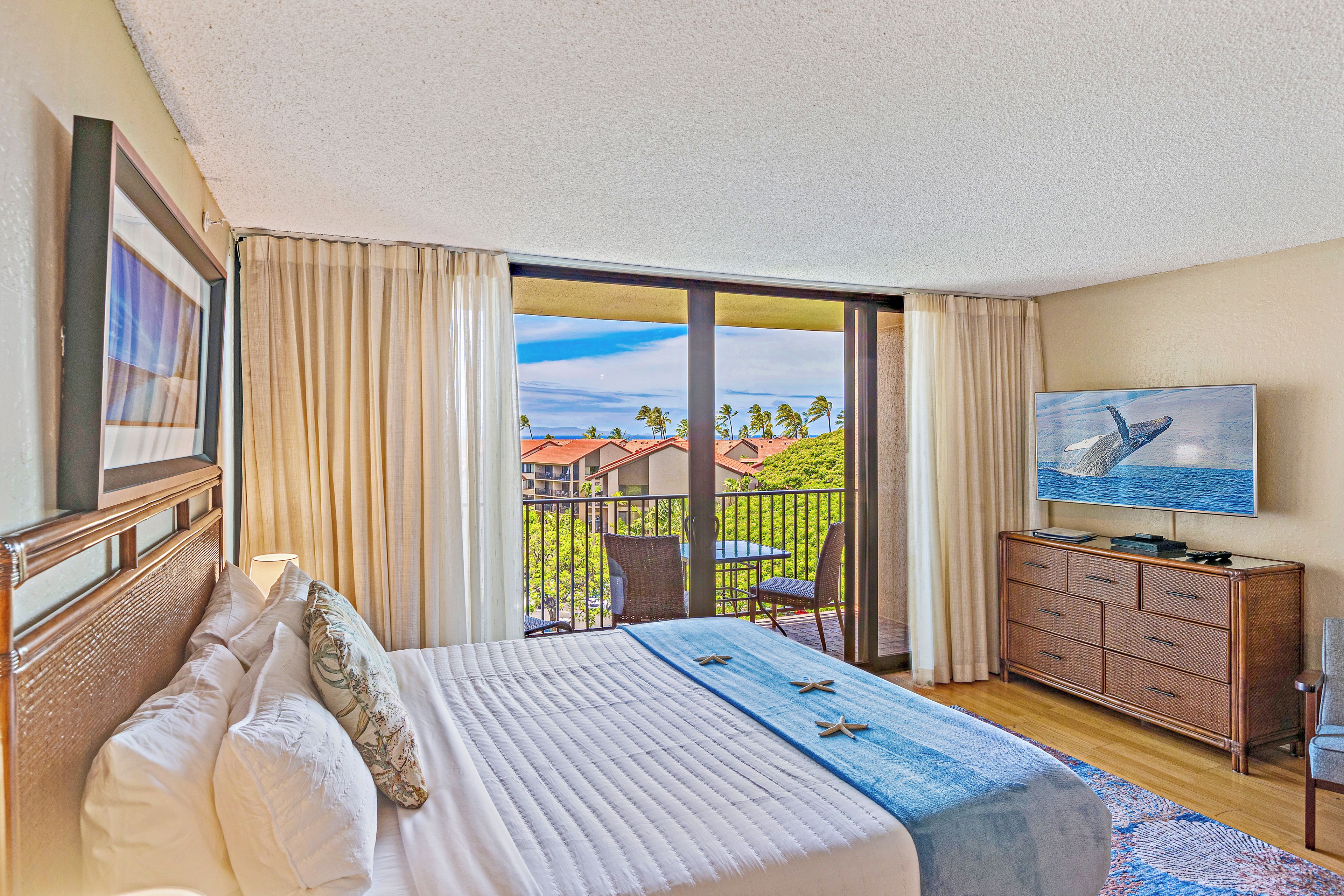 Habitación de hotel con cama, televisor, cómoda y balcón con vistas a palmeras y casas.