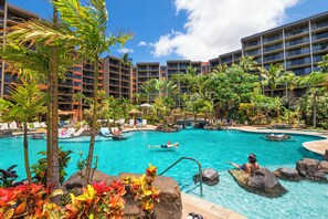 Zona de piscina hotelera con varios niveles de balcones, rodeada de plantas tropicales y un cielo azul despejado.