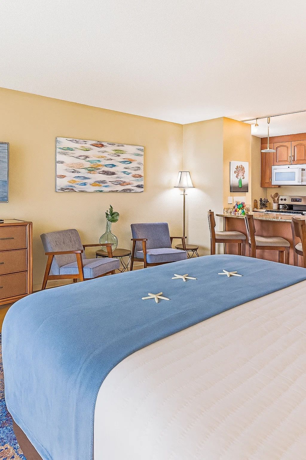 A hotel room with a bed, two chairs, a desk, a lamp, a microwave, and a painting on the wall.