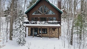 Hirsimökki, jonka perustuksena on kivimuuri, siinä on suuret ikkunat ja parveke, jonka kaiteet ovat lumen peitossa.