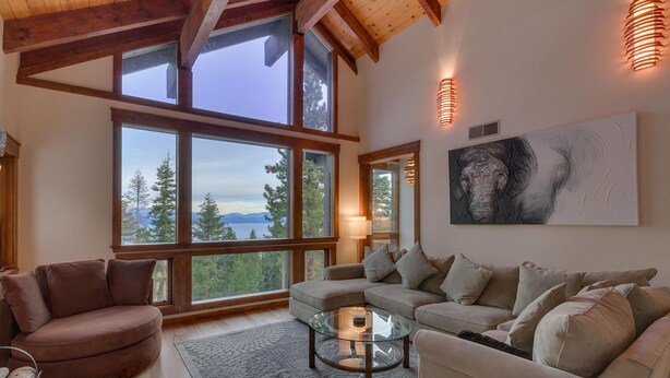 Un salon spacieux doté de grandes fenêtres offrant une vue sur les arbres et les montagnes, un canapé moderne, une table basse en verre et une cheminée.