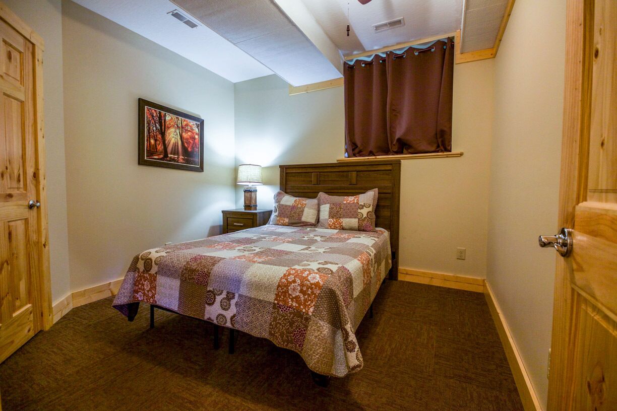A bedroom with a wooden headboard, a quilted bedspread, a nightstand with a lamp, and a window with curtains.