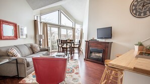 A spacious living room with a large window, a fireplace, a dining area, and a TV mounted on the wall.