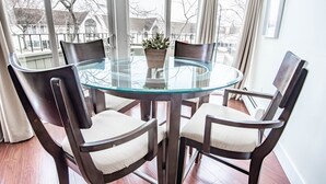 A round glass table with four dark wooden chairs, a potted plant, and a view of a balcony with a railing and trees.