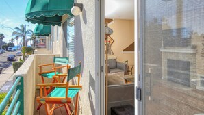 Un balcón con toldo verde, un espacio de estar con sofá y sillones, y vista a la calle con palmeras y coches estacionados.