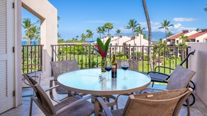 Une table ronde en verre, entourée de quatre chaises, avec deux bouteilles de vin et deux verres à vin, est placée sur un balcon qui offre une vue imprenable sur un paysage tropical, avec des palmiers et une chaîne de montagnes en arrière-plan.