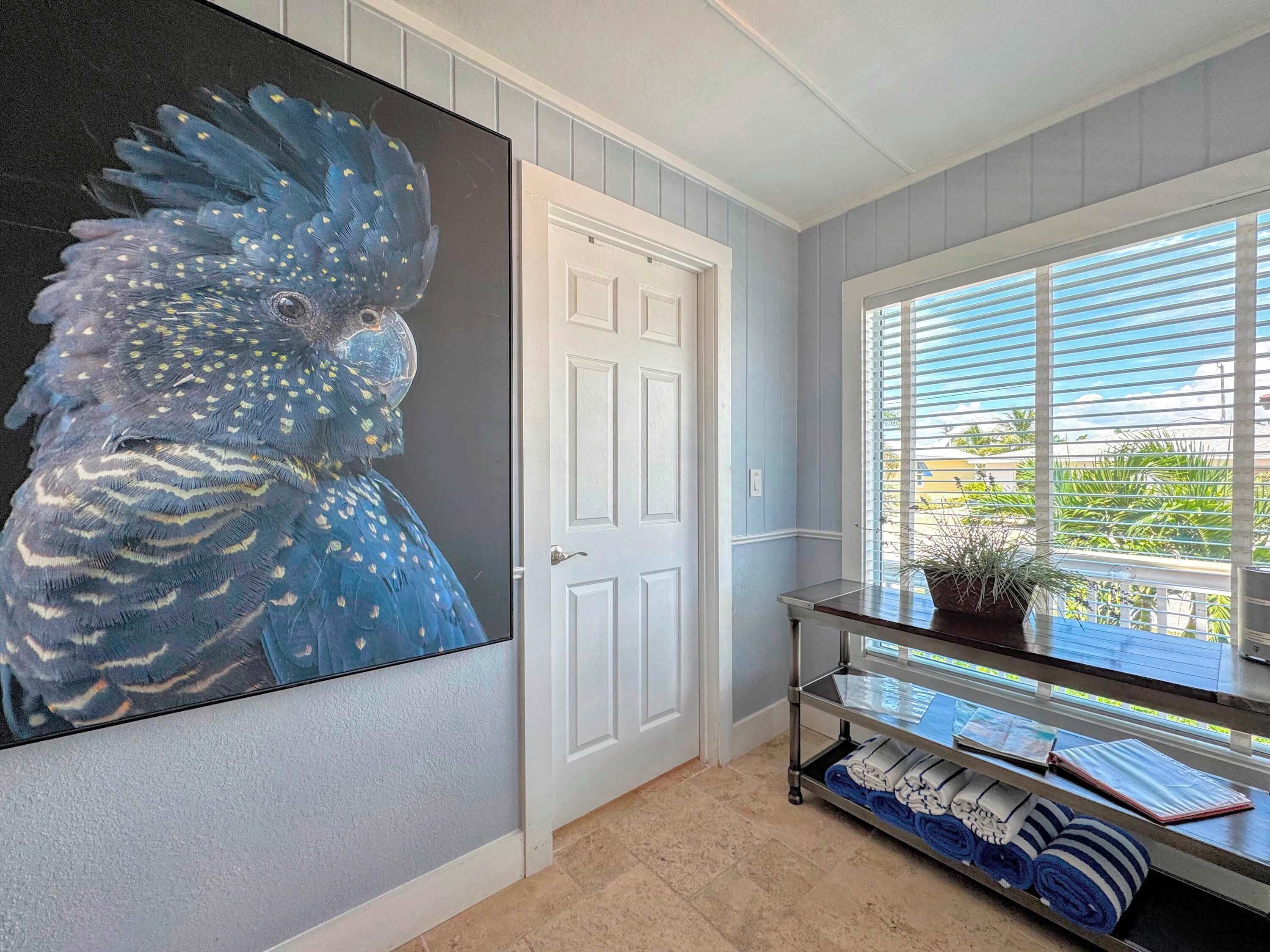 A hallway with a large painting of a bird, a white door, a window with blinds, and a shelf with towels.