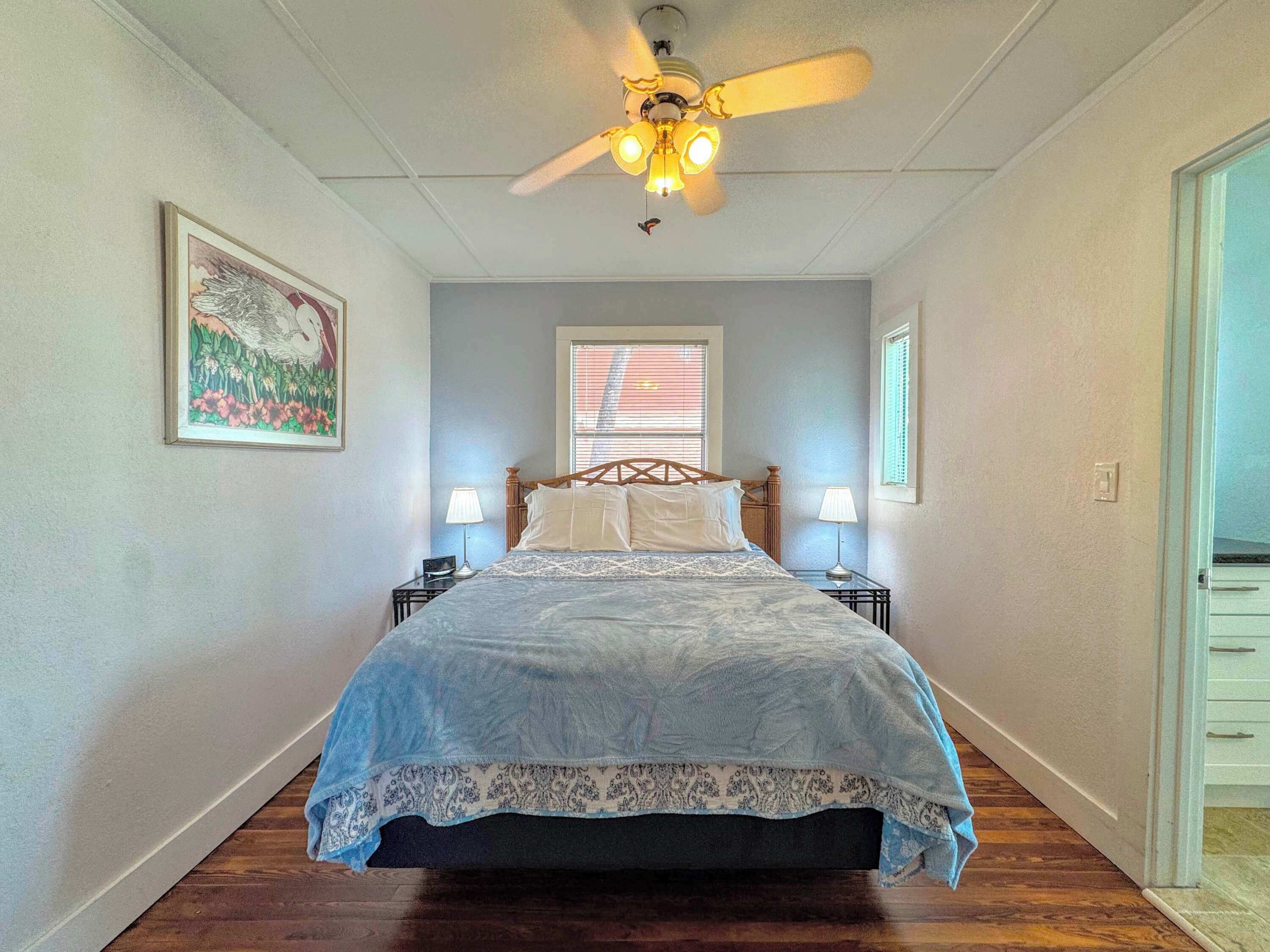 A bedroom with a bed, two bedside tables, a ceiling fan, a window with blinds, and a door.