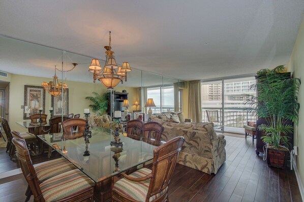 A spacious living room with a large glass dining table surrounded by wooden chairs, a patterned sofa, and a large window offering a view of the city.