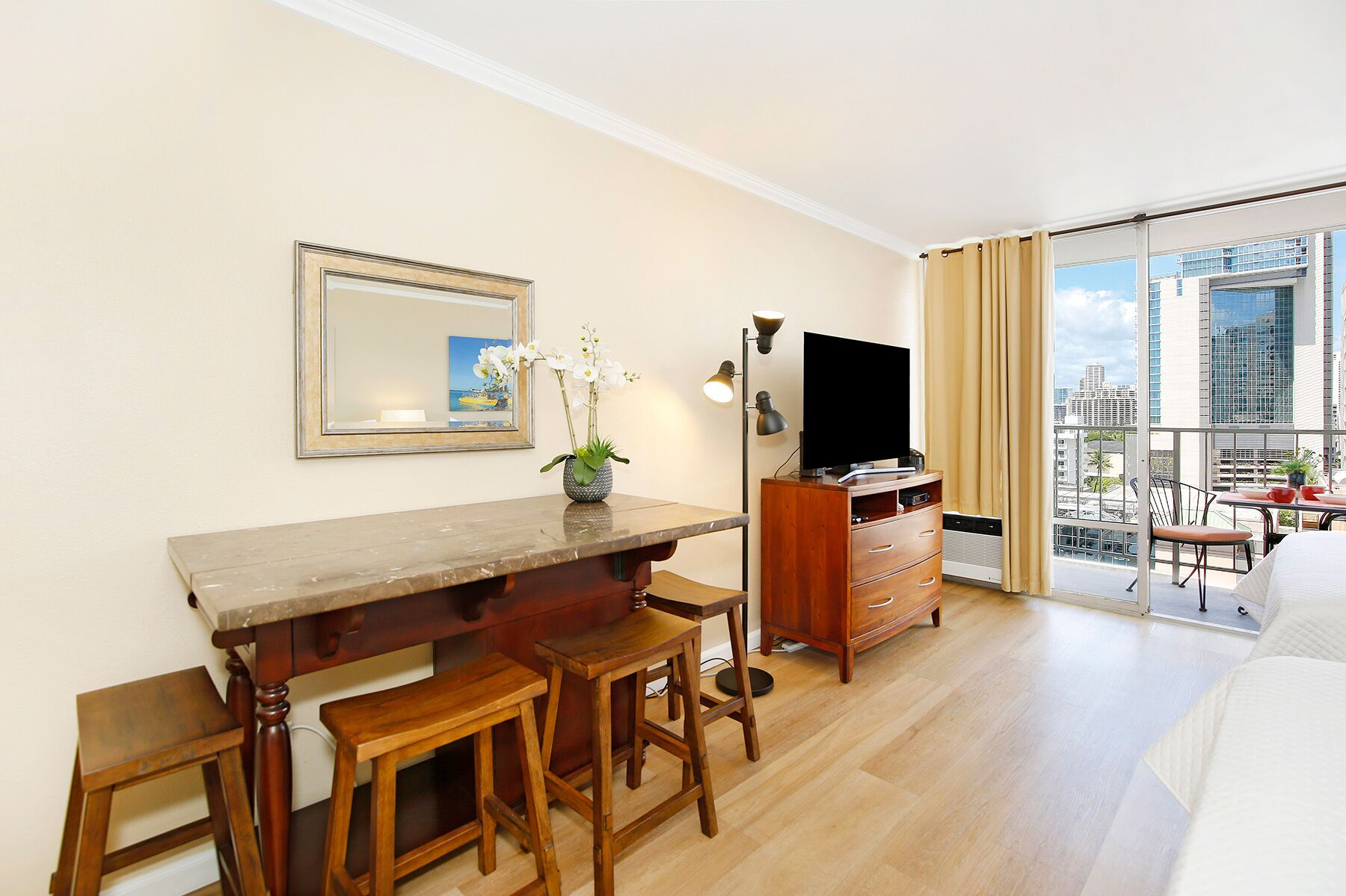 A modern hotel room with a wooden desk, a flat-screen TV, a large window with a city view, and a balcony with outdoor seating.