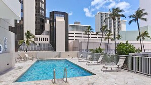 Piscina no terraço com água azul cristalina, cercada por espreguiçadeiras e palmeiras, com prédios modernos ao fundo.
