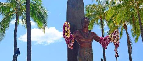 Une statue représentant un homme tenant une planche de surf, entouré de palmiers et de parasols de plage.