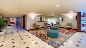 Um hall de hotel com piso ladrilhado, um tapete colorido, cadeiras de vime e obras de arte emolduradas nas paredes.