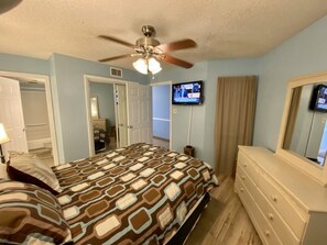 A bedroom with a bed, dresser, ceiling fan, TV, and a door leading to another room.
