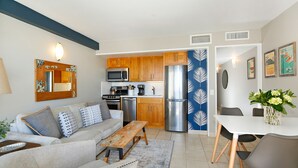 Ein modernes Wohnzimmer mit Sofa, Couchtisch und Esstisch. Der Küchenbereich umfasst Holzschränke, eine Mikrowelle und einen Kühlschrank.