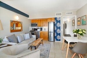 A modern living room with a sofa, coffee table, and dining table. The kitchen area includes wooden cabinets, a microwave, and a refrigerator.