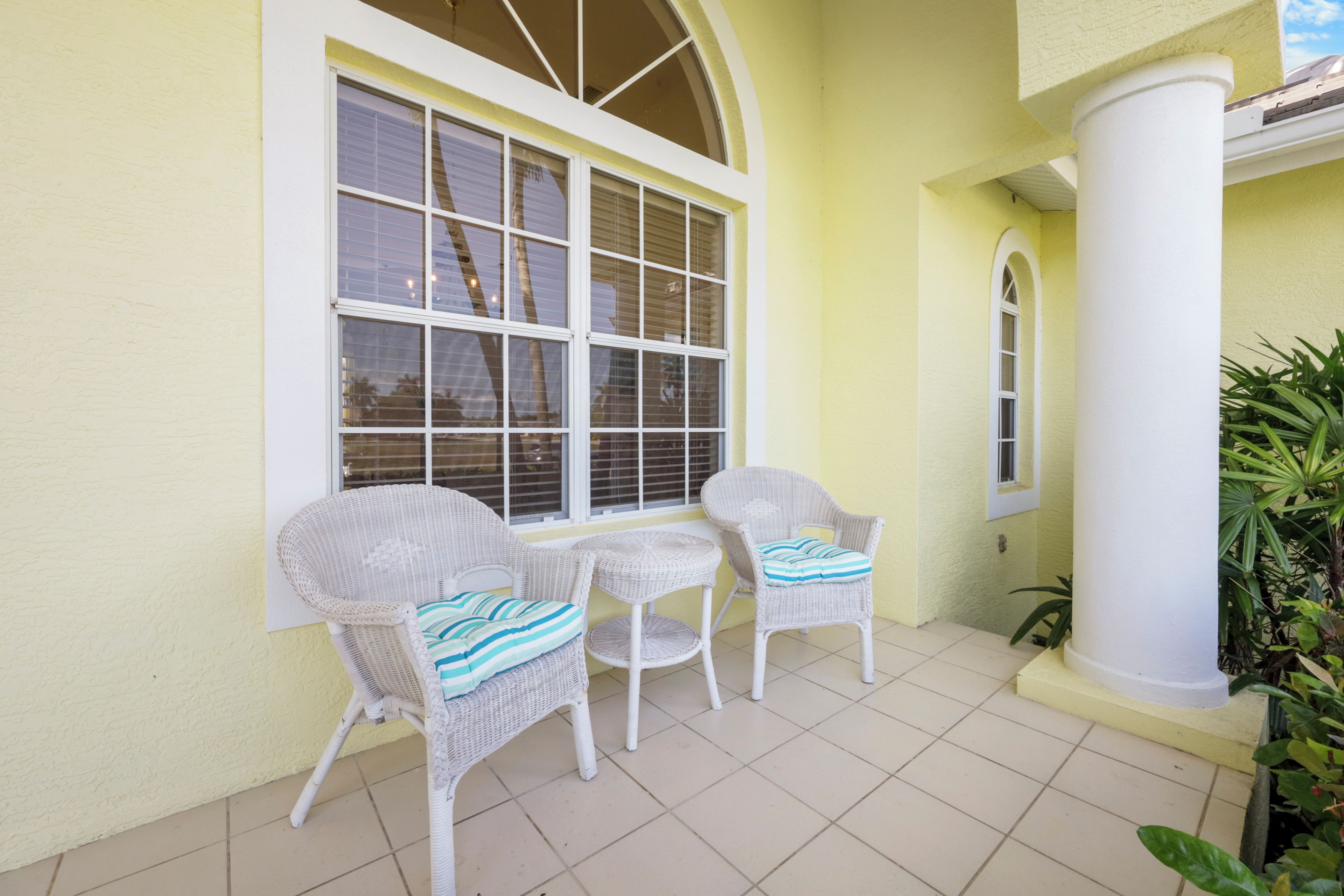 A patio with two wicker chairs and a small table, a large window with blinds, and a white column.