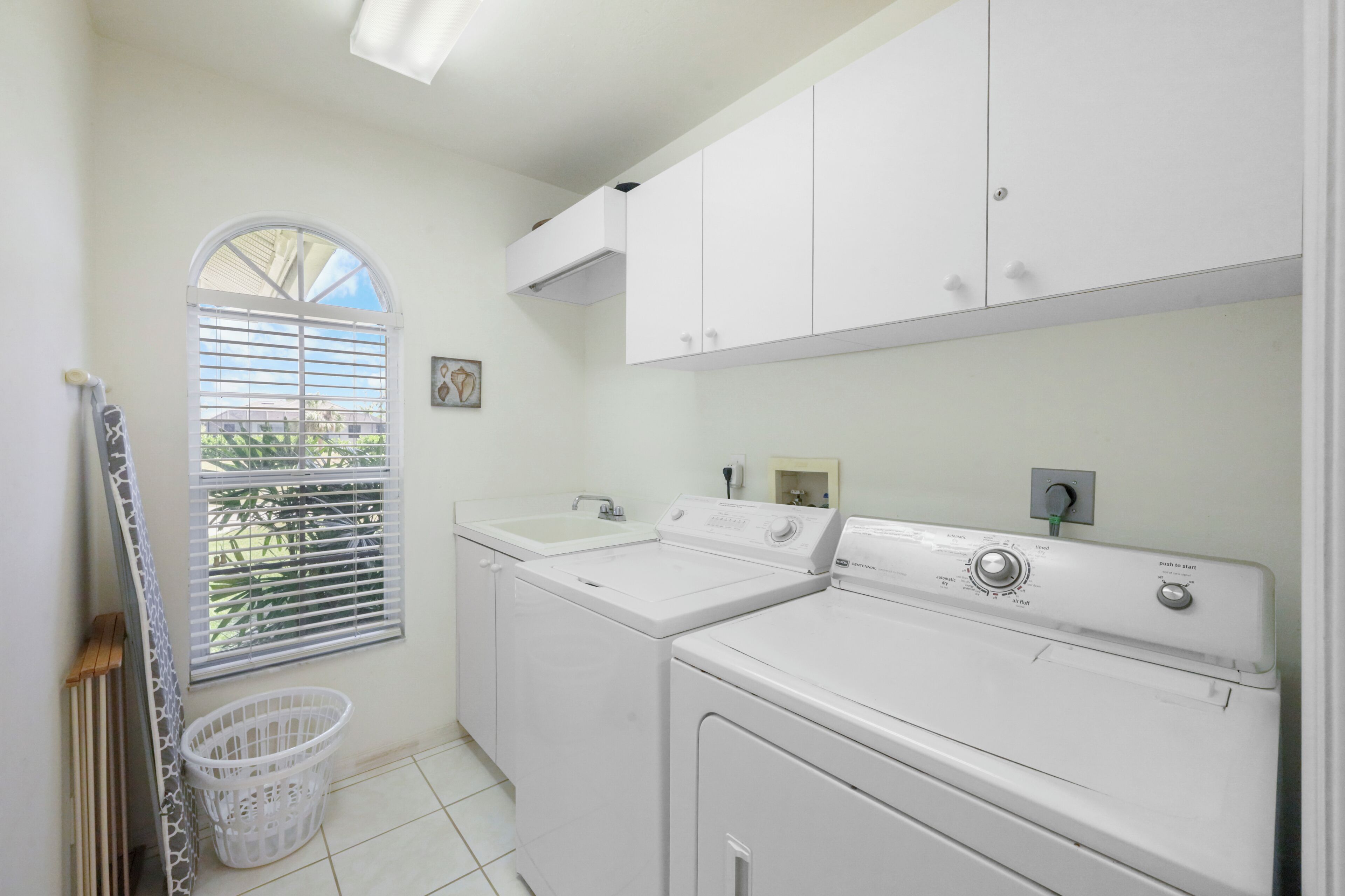 A laundry room with two white washing machines, a sink, and a window with blinds.