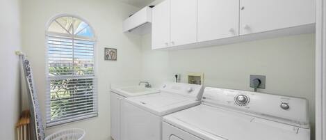 A laundry room with two white washing machines, a sink, and a window with blinds.