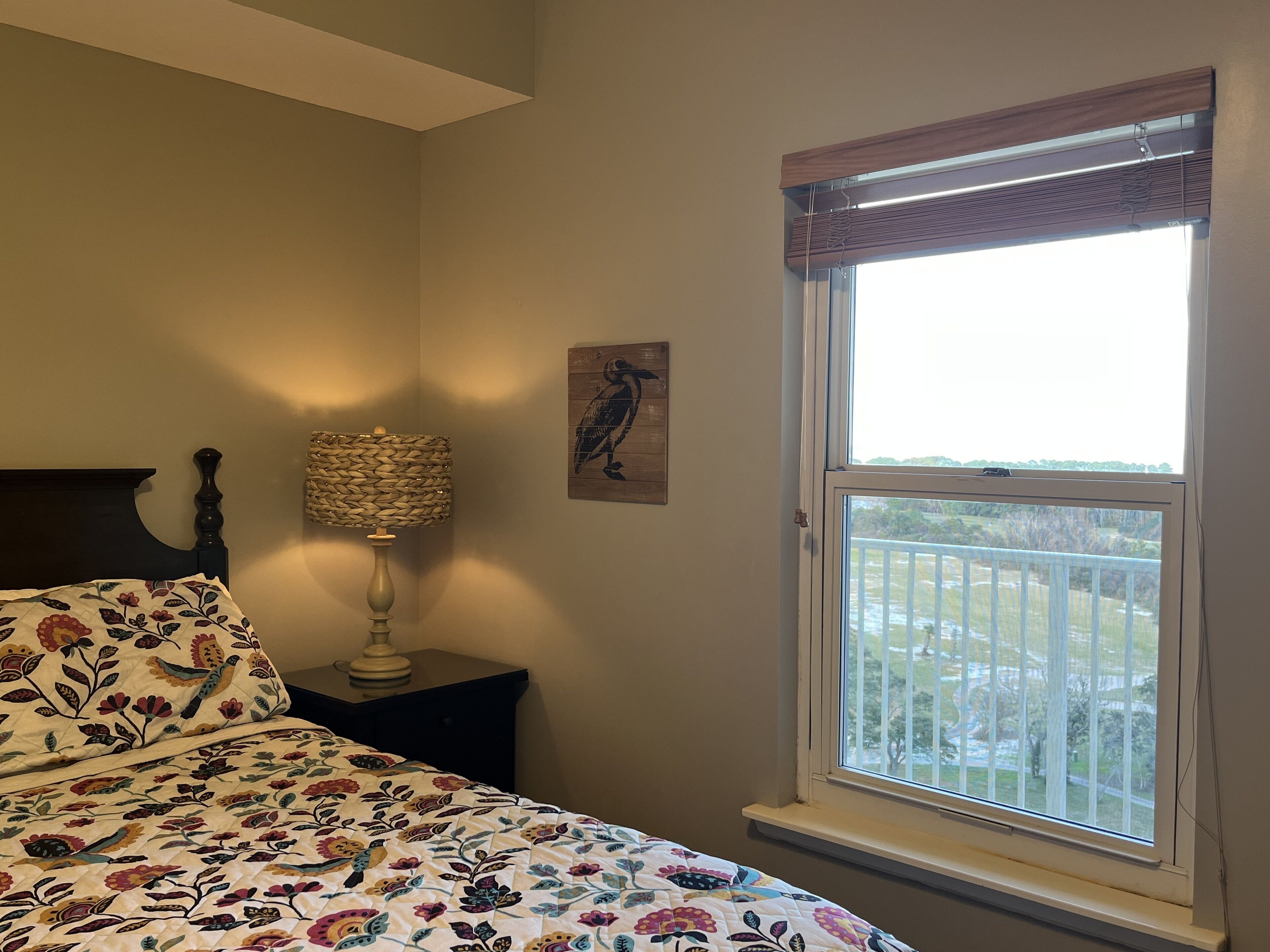 A bedroom with a bed, a nightstand, a lamp, a window with a view of a balcony, and a wall-mounted artwork.