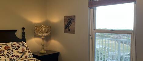 A bedroom with a bed, a nightstand, a lamp, a window with a view of a balcony, and a wall-mounted artwork.