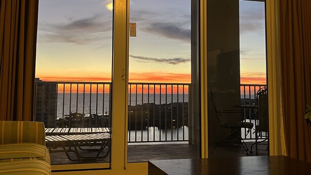 Una habitación con puerta corrediza de vidrio que da a un balcón con vista a la puesta de sol sobre el agua.