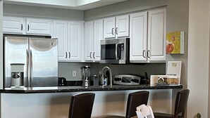 Una cocina moderna con armarios blancos, electrodomésticos de acero inoxidable y encimera negra. Hay tres sillas marrón oscuro alrededor de una mesa pequeña.
