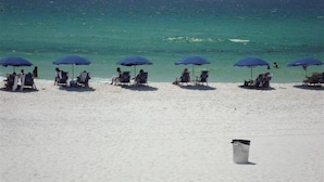 Una playa con hileras de sillas y sombrillas, un bote de basura blanco, y el océano al fondo.