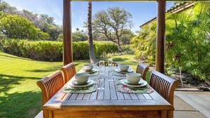 A wooden table set for four with bowls and wine glasses, under a wooden pergola, surrounded by lush greenery and a clear sky. - Private luxury home with high ceilings and room for 6 (Kamuela)