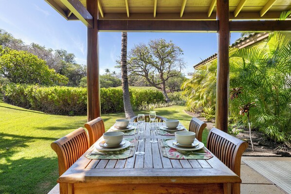 A wooden table set for four with bowls and wine glasses, under a wooden pergola, surrounded by lush greenery and a clear sky.