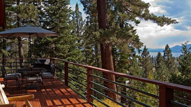 Una terraza de madera con mobiliario exterior, una mesa y sillas, con vistas a un bosque y un lago.