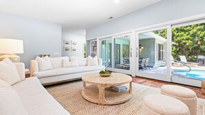 A modern living room with a white sofa, a round wooden coffee table, and a large glass door leading to an outdoor pool area.