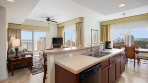 A modern kitchen with a dishwasher, a dining area with a table and chairs, a flat-screen TV, and a balcony with a view of the city. - Lockout Style ~ Gulf Views ~ Balcony ~ Resort Pool (Miramar Beach)