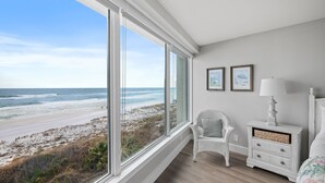 A bedroom with a large window overlooking a beach, a wicker chair, a bedside table with a lamp, and two framed pictures on the wall. - Beachfront ~ Private Balcony ~ Recent Updates (Miramar Beach)