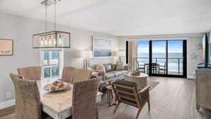 A spacious living room with a large window overlooking the ocean, a dining area with wicker chairs, and a modern chandelier. - Beachfront ~ Private Balcony ~ Recent Updates (Miramar Beach)