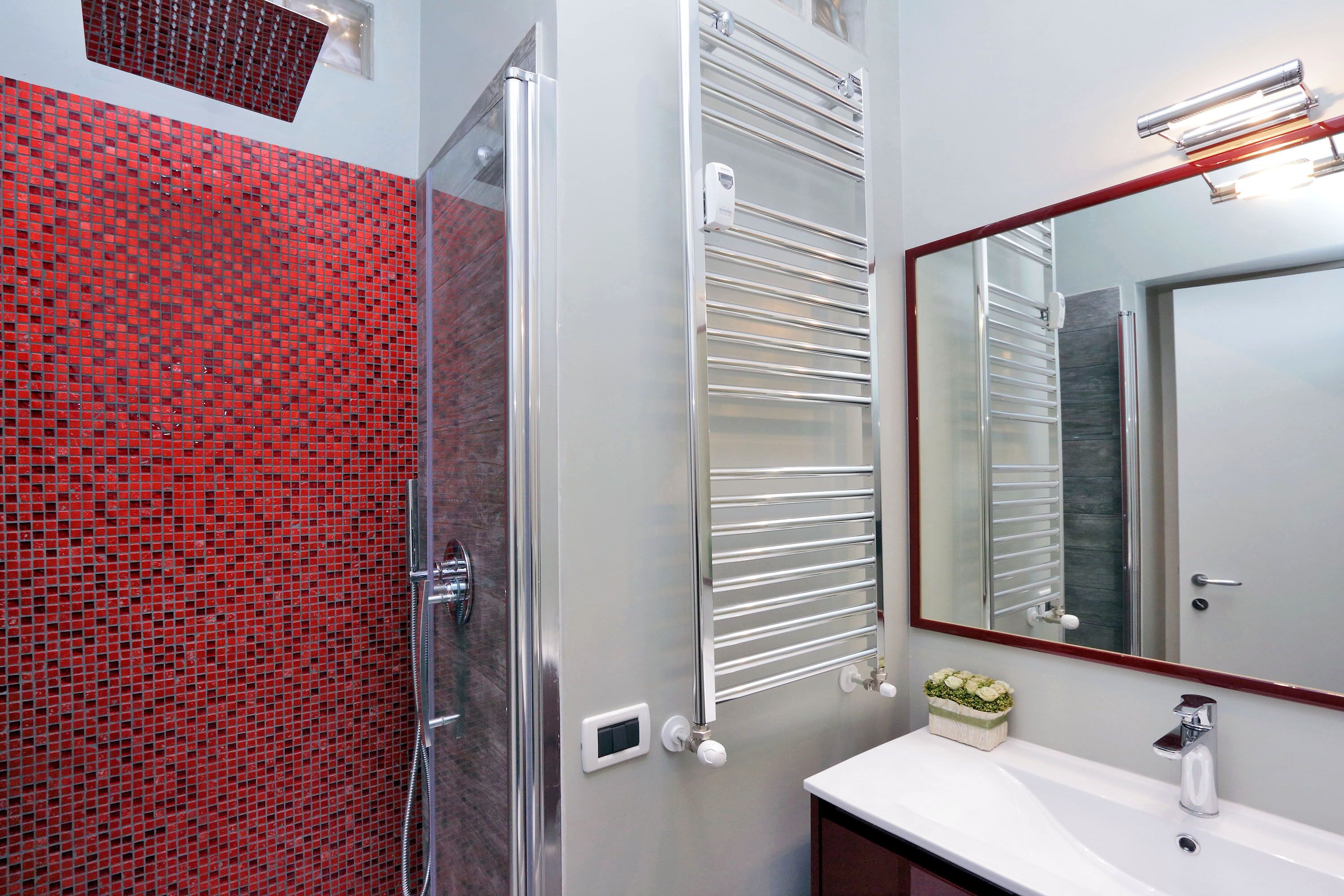 Ein Badezimmer mit roter Mosaikwand, einer Glasdusche, einem weißen Waschbecken und einem Spiegel mit rotem Rahmen.