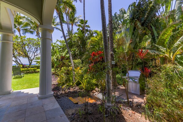 A tropical garden with palm trees, a barbecue grill, and a view of the ocean. - Peaceful Oceanfront Studio 423 LS (Lahaina)