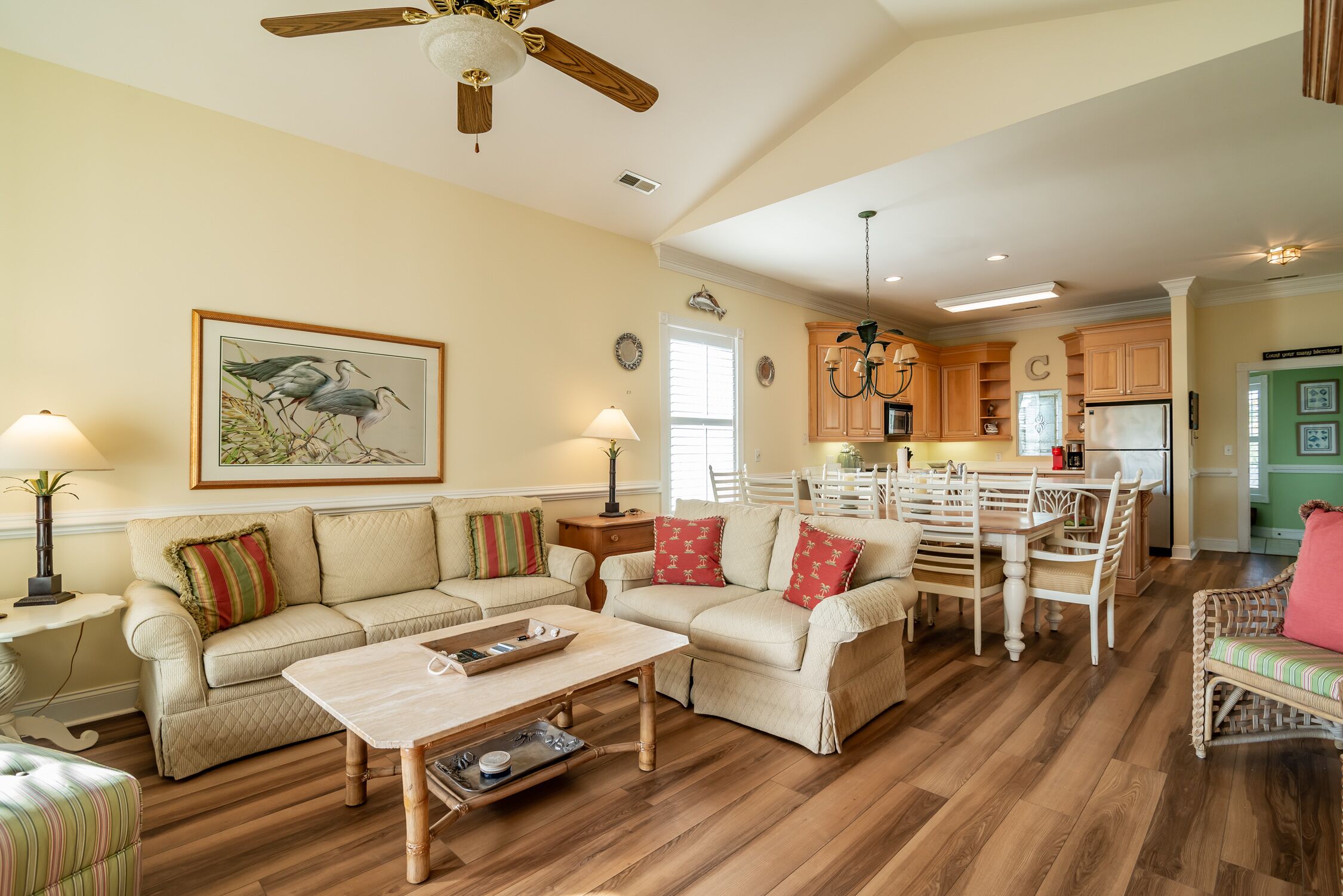 Ein geräumiges Wohnzimmer mit Holzboden, zwei beigefarbene Sofas mit roten und grünen Kissen, einem Holztisch und einem Essbereich mit Tisch und Stühlen.