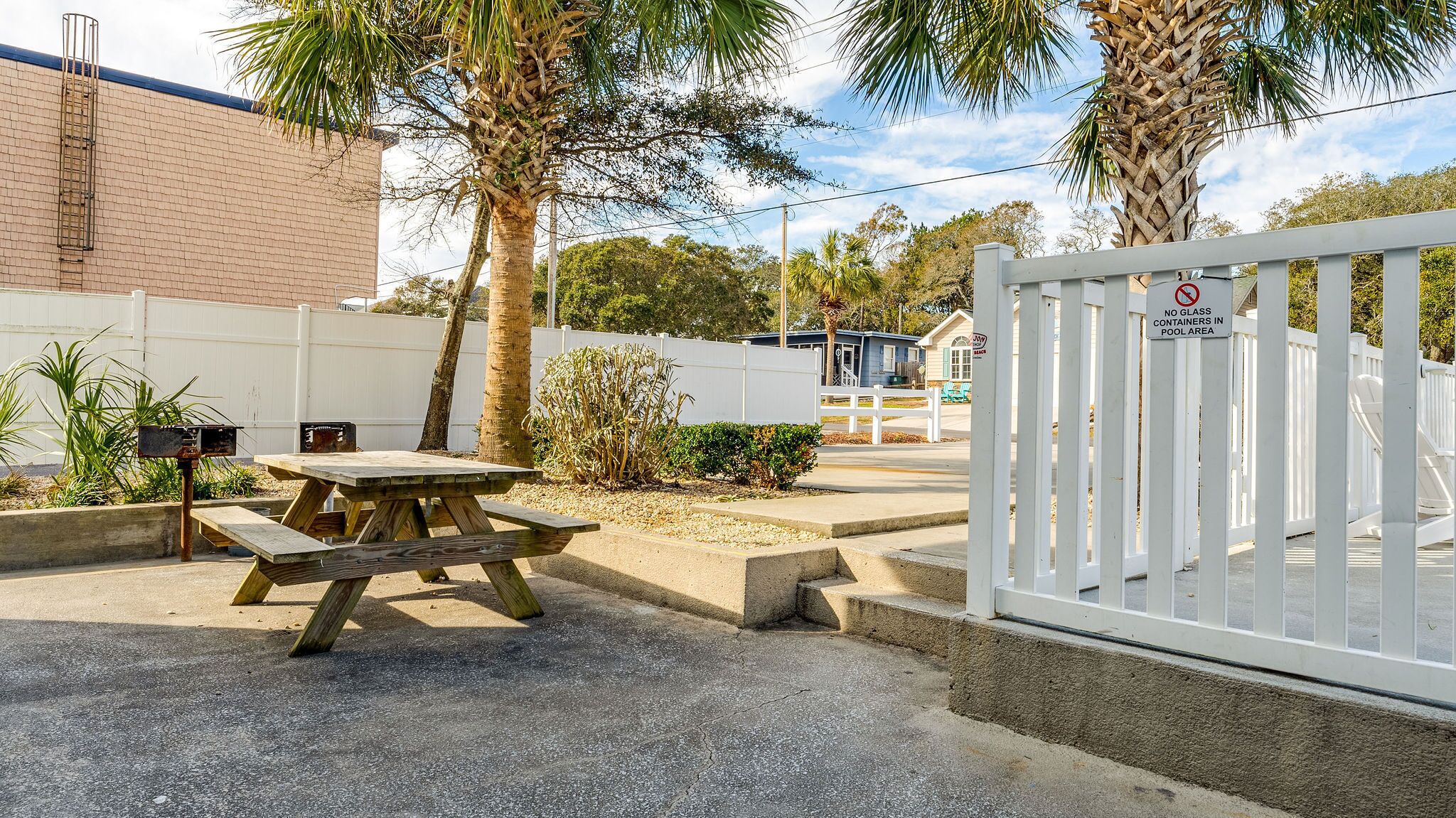 Een picknicktafel met bankjes, een wit hek en een bordje dat glazen containers bij het zwembad verbiedt.