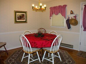 Comedor con mantel rojo, sillas blancas, candelabro, un cuadro enmarcado y una ventana con cortinas.