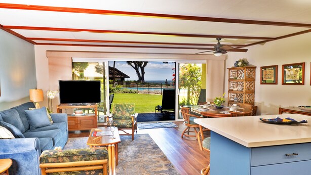 A spacious living area with a sofa, chairs, a coffee table, and a TV. There's a large window with a view of a lawn and a building.