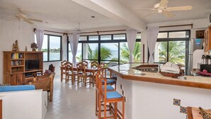A spacious living area with a large window overlooking a body of water, a wooden bar area, and a television on a wooden stand. - Spacious with beautiful upper level views of Half Moon Bay! (Akumal)