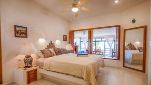 A bedroom with a bed, nightstands, a ceiling fan, a large window, and a mirror. - Spacious with beautiful upper level views of Half Moon Bay! (Akumal)
