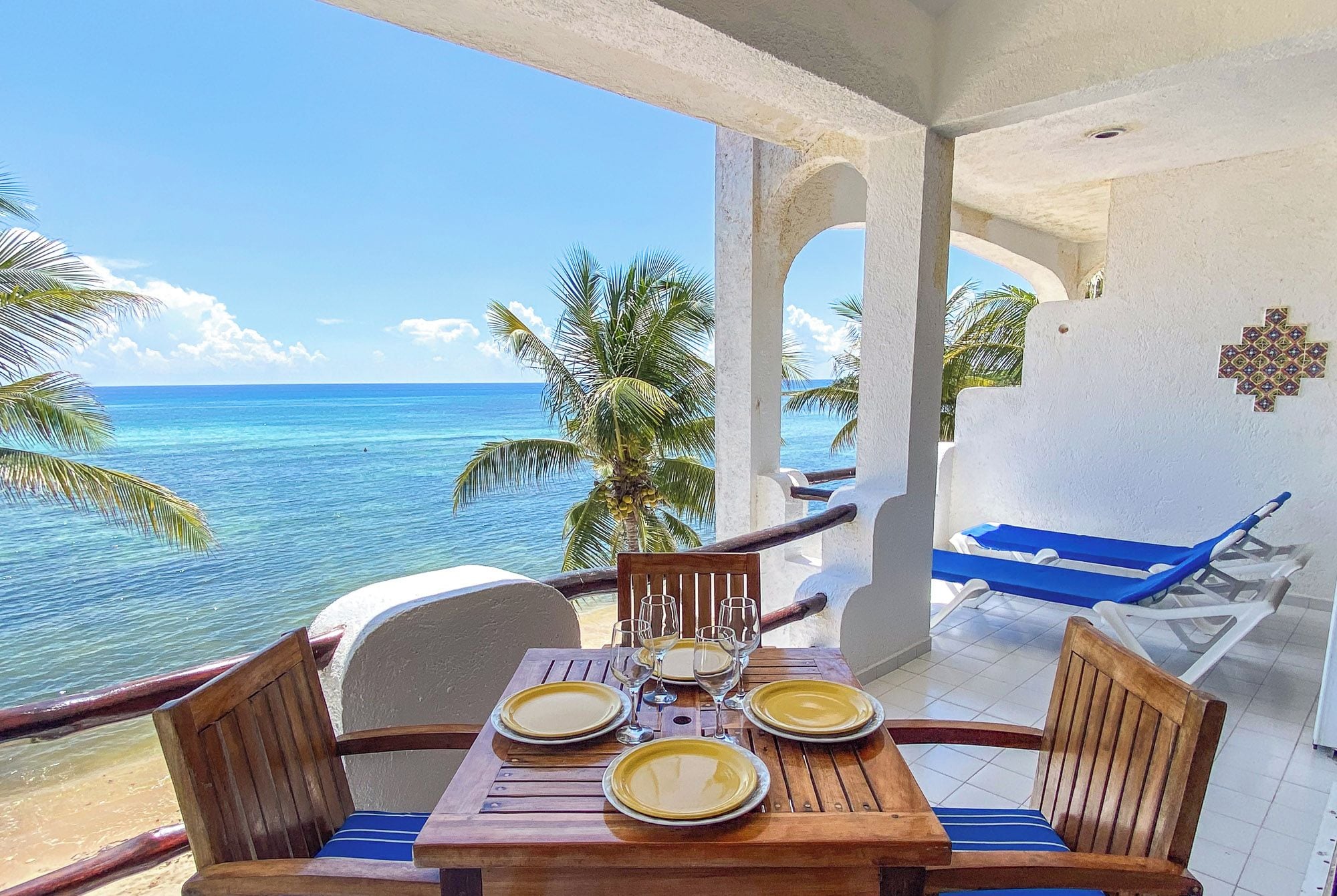 A wooden table set for four with plates and glasses, overlooking a beach with palm trees and the ocean.