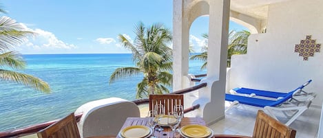 A wooden table set for four with plates and glasses, overlooking a beach with palm trees and the ocean.