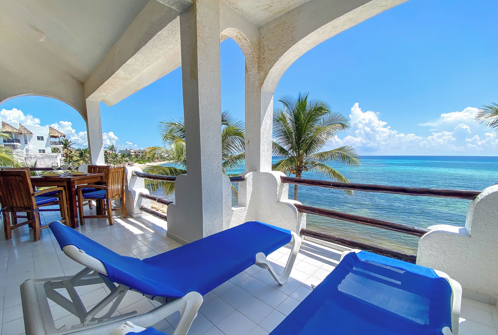 A balcony with blue lounge chairs, a wooden table with chairs, and a view of the ocean.