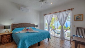 Ein Hotelzimmer mit Bett, Deckenventilator, Fenster mit Vorhängen und Blick auf den Strand sowie ein Stuhl am Fenster.